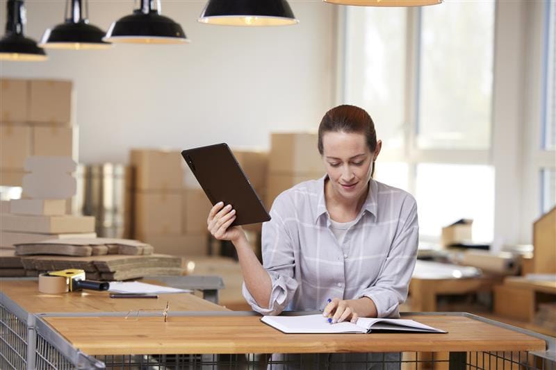 Frau in einem modernen Büro, die mit einem Tablet in der Hand Notizen in ein Notizbuch macht, umgeben von Kartons und Büromaterialien. Ideal für Themen wie Produktivität, Büroorganisation und digitales Arbeiten.