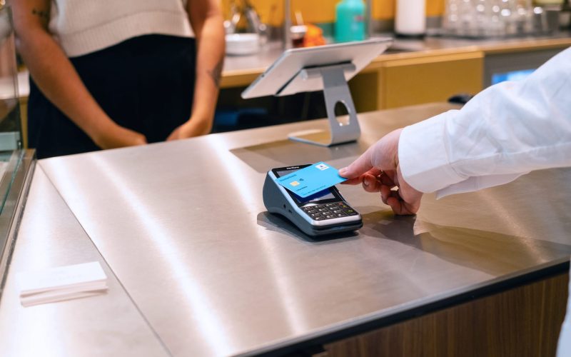 Zahlungsvorgang an der Kasse mit einem Kartenlesegerät in einem modernen Café, während eine Person eine Kreditkarte vor das Gerät hält. Im Hintergrund ist eine weitere Person zu sehen, die auf den Zahlungsvorgang wartet.
