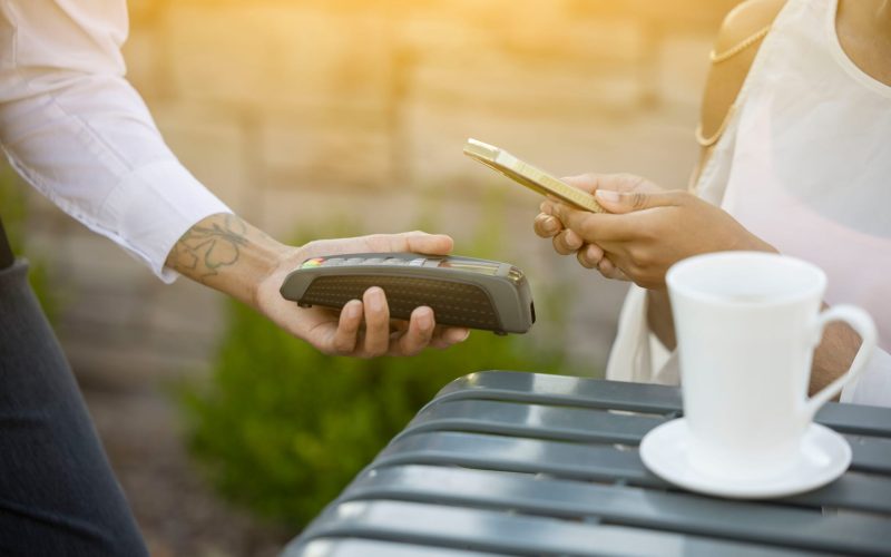 Zahlungsvorgang in einem Café, bei dem ein Kunde sein Smartphone zur Bezahlung nutzt, während ein Mitarbeiter das Kartenlesegerät überreicht. Im Hintergrund ist eine Tasse Kaffee auf dem Tisch sichtbar.